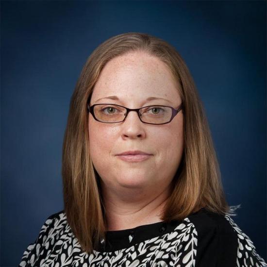 A woman with straight, shoulder-length light brown hair, wearing glasses and a black-and-white patterned blouse, is set against a dark blue background.